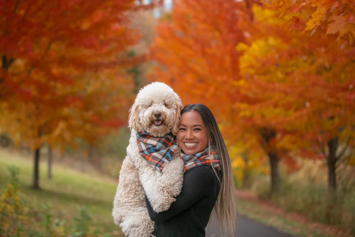 Woman holding her doodle in a plaid bandana under orange autumn maples