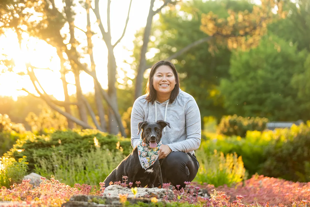 Woman crouching with her dog in a bandana in a garden at sunrise